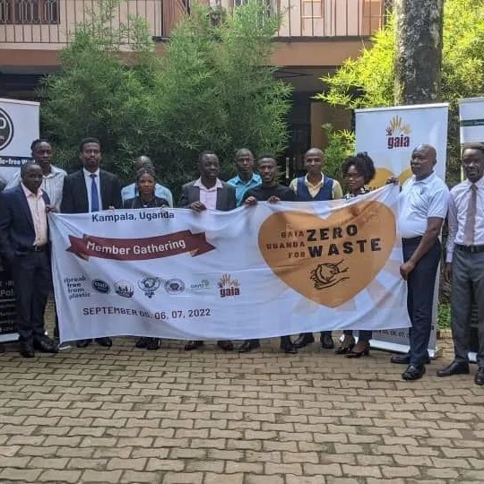 A picture of the Uganda members at their first members meeting in Kampala. They are holding a banner that reads “Member Gathering“ with a yellow heart that reads “GAIA Uganda for Zero Waste“ and a drawing of hands holding the Earth
