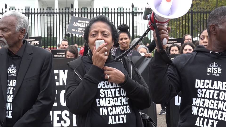 A photo of Sharon Levigne in front of the gates of the White House speaking into a megaphone. She and other participants are wearing black shirts with white text that read: “RISE ST. JAMES: DECLARE A STATE OF EMERGENCY IN CANCER ALLEY.“