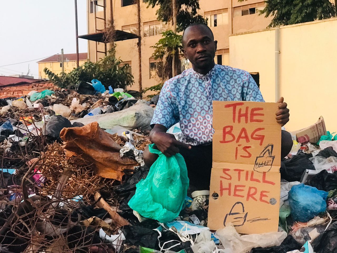 Nirere Sadrach from End Plastic Pollution, Uganda standing outside amidst a pile of plastic waste, holding a sign that says “THE BAG IS STILL HERE“