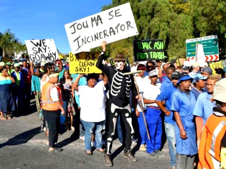 A photo of community members during an anti-incineration protest in Wellington, South Africa. In the foreground, a person wearing a skeleton outfit with a sign that reads, “Jou Ma Se Incinerator.“ Other signs read “We Say: Go Away,“ “Toxic Ash From Trash,“ and “Bye Bye Clean Sky.“