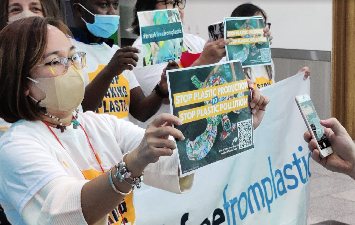 A photo taken at the 7IMDC of a member holding a sign that reads, “STOP PLASTIC PRODUCTION. STOP PLASTIC POLLUTION,“ over a photo of plastic waste. In the background, other members are seen holding similar signs as well as a #breakfreefromplastic banner. Members are wearing shirts that read “Stop Making Plastic.“